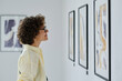 © AnnaStills - Young woman in eyeglasses standing in front of pictures on the wall, examining the art and smiling