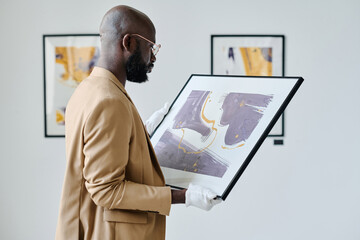 African worker examining picture in his hands during preparation for exhibition at art gallery