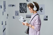 © AnnaStills - Young woman in wireless headphones reading booklet while standing at art gallery