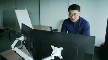 Concentrated asian programmer, financial analyst or trader working at computer desk in office. Thoughtful it specialist employee in the office is thinking about solving problems looks at the monitor