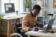 Portrait of black young man with disability working in office and using laptop, accessible workplace concept