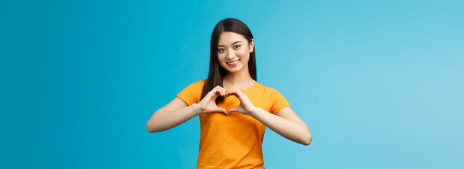 Wall Mural - Lovely caring attractive asian girlfriend show heart sign cherish and value relationship, smiling broadly supportive, stand blue background wear yellow t-shirt, promote peace and happiness