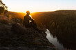 © Francis - A male hiker sitting on a rocky cliff edge at sunset, looking over a vast forest valley and river. Serene outdoor adventure, wilderness exploration, and golden hour landscape photography.