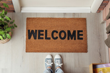 Woman standing near door mat with word Welcome on wooden floor in hall, top view