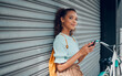 © Kay Abrahams/peopleimages.com - City lifestyle, girl with phone and bike standing at wall thinking before writing text. Happy female student, bicycle and smartphone outside street, communication, 5g and sustainable urban transport.