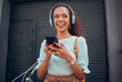 © Kay Abrahams/peopleimages.com - Black woman, smile and phone for social media in the city with headphones in joy for 5G connection in the outdoors. Happy African female student smiling for technology or internet on smartphone