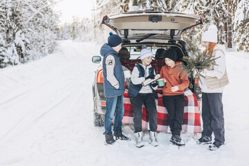 Wall Mural - Four friends teenagers boys and girl with thermos sitting in trunk of car decorated for Christmas and New year. Winter picnic in snowy forest. Road trip and local travel. Friends hanging out together