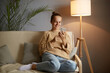 © sementsova321 - Image of smiling delighted young adult woman wearing casual clothing sitting on sofa and using mobile phone for checking social networks, chatting with friends.