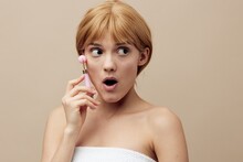 A Joyful, Funny Woman, Blonde, With Her Hair Pinned Back, Stands On A Beige Background Wrapped In A Towel And Does A Facial Massage With A Pink Roller, Her Mouth Wide Open. Skin Care Topics