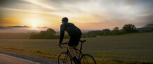  Cyclist Riding His Bicycle On An Empty Rural Road At Dawn, Epic Sunrise In The Background