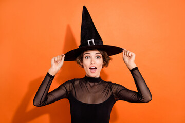 Wall Mural - Photo of sexy impressed young lady wear black witch dress hands cap open mouth isolated orange color background