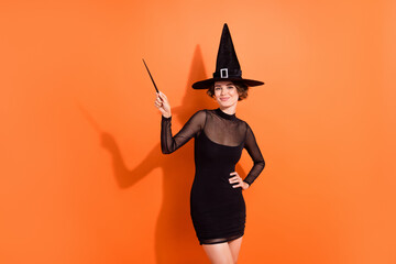 Wall Mural - Photo of cute young woman point wooden wand hogwarts theme event dressed trendy black halloween witch look isolated on orange background