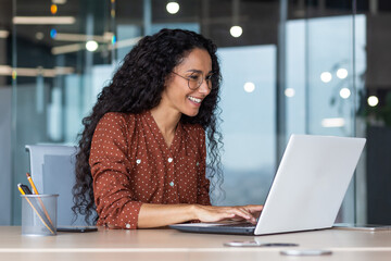 Wall Mural - Young and happy hispanic woman working in modern office using laptop, business woman smiling and happy in glasses and curly hair.