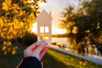 Wall Mural - The girl holds a paper house in her hand against the background of the sunset sky. The sun is setting over the horizon. The concept of your own home, personal space and mortgage