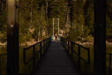 A Caucasian Man Wearing A Beanie And A Brown Jacket With A Backpack Crossing A Bridge Over A River In The Forest.