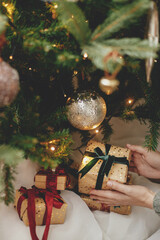 Wall Mural - Stylish christmas gift in hands under christmas tree with lights. Merry Christmas and Happy Holidays! Woman in cozy sweater putting wrapped christmas present in atmospheric festive room