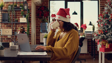 Startup Employee Writing Report On Laptop During Christmas Eve, Working In Office Decorated With Seasonal Ornaments And Lights. Festive Woman With Santa Hat Sending Email During Xmas Holiday.