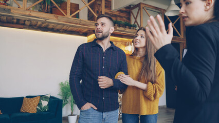 Wall Mural - Confident housing agent is showing spacious modern house with beautiful interior to happy young couple wife and husband, people are smiling and talking looking around.