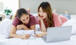 © Bangkok Click Studio - Millennial Asian young happy cheerful female teenager LGBTQ lesbian lover couple laying down together on bed and pillow learning studying online via laptop computer and writing short note on notebook