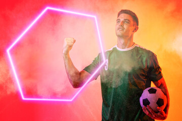 Wall Mural - Caucasian male player holding soccer ball clenching fist over illuminated hexagon on neon background