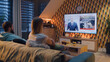 © Framestock - Couple sitting on couch in living room with snacks, watching TV news about military exercises, NATO training, armed forces. Man and woman spending evening at home. TV live broadcasting.