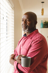 Wall Mural - Vertical image of thoughtful african american senior man standing by window with coffee smiling