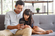 © aekkorn - Cute asian girl help young father show something on smartphone making selfie together, smart daughter and dad sit on couch hold mobile phone take family picture on moblie phone.