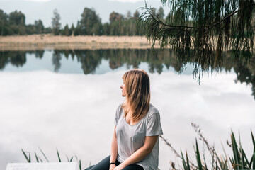 Canvas Print - blonde caucasian girl in gray t-shirt and black pants in profile thinking and admiring the beauty of the surroundings sitting on the shore of the lake with transparent waters near the forest and