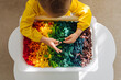 Child playing with sensory bin with dried pasta in rainbow colors. Dyed pasta for play and craft activities. Montessori material. Sensory play and learning colors activity for kids.