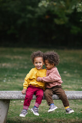black and asian twins hugging on bench in park while smiling