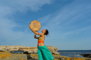 A woman in boho style clothes with a shamanic ritual tambourine on the rocks on the seashore on a clear sunny day. The concept of freedom of mind and body in nature