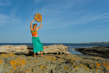 A woman in boho style clothes with a shamanic ritual tambourine on the rocks on the seashore on a clear sunny day. The concept of freedom of mind and body in nature