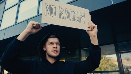 serious young man in hoodie holding placard with no racism lettering outdoors.