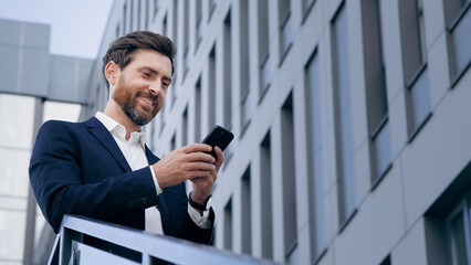 Stylish handsome businessman holding phone reading email stands on street smiling writes message communicates in social network pensive man browses goods in online store using mobile app on smartphone