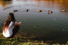 Young Woman Feeding Ducks While Sitting By The Lake During Sunny Weather In Autumn