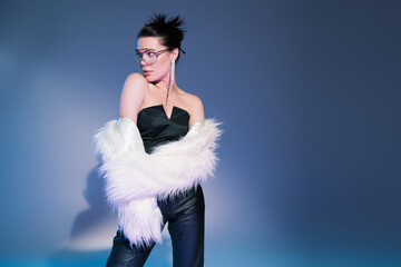 glamour model in black corset with leather pants looking away while posing in white faux fur jacket on blue background.