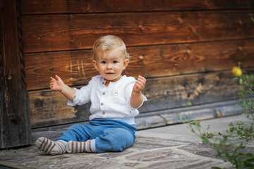 blond one year old baby boy in chic white  shirt looking serious & focused into the camera outside of a rustic wooden hut  while clapping hands and making hand signs