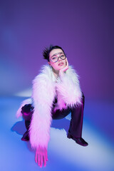 Wall Mural - trendy woman in transparent eyeglasses and white fluffy jacket looking at camera on purple background.