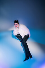 Wall Mural - high angle view of fashionable woman in white furry jacket looking at camera while posing on blue background.