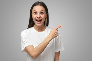 Wall Mural - Young surprised woman in white t-shirt pointing right shouting WOW on gray background