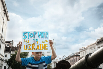 Wall Mural - Boy holding poster in colors of national flag with words Stop War In Ukraine near broken military tank on city street, closeup