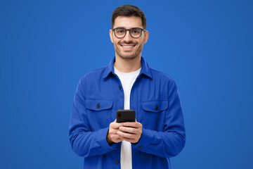 Young modern man in casual blue shirt and glasses, holding smart phone in hands, looking at camera