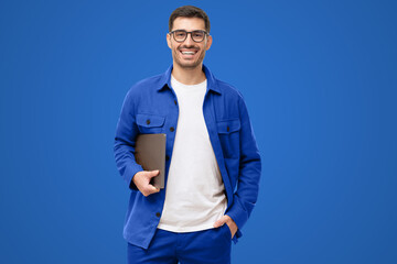 Wall Mural - Young smiling modern man or male teacher holding laptop, isolated on blue
