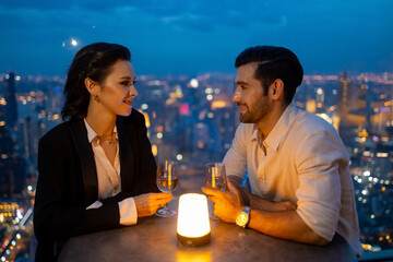 Caucasian couple celebrating holiday event at luxury skyscraper rooftop bar at night. People enjoy city nightlife having dinner and drinking wine at outdoor rooftop restaurant on summer vacation.