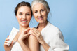 Mother and daughter holding skin cream and smiling. Two beautiful women of different ages and generations together on a blue background.