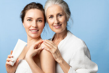 Wall Mural - Mother and daughter holding skin cream and smiling. Two beautiful women of different ages and generations together on a blue background.