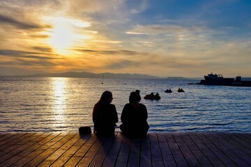 Two girls sitting on the pier by the sea coast on a sunset