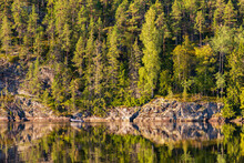 .A Rocky Slope Overgrown With Dense Green Trees Is Reflected In The Still Lake Water, Karelia, Russia