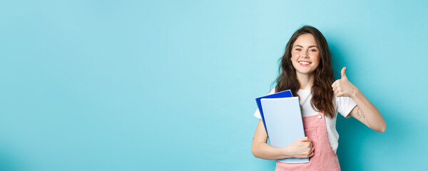 Young woman attend courses, girl student studying, holding notebooks and showing thumb up in approval, recommending company, standing over blue background