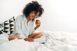 © Daniel - Smiling young mixed race woman reading journal notes on bed drinking some tea. Copy space.